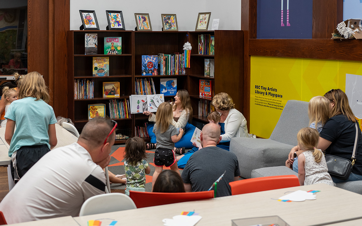 Parents and kids gather by the bookshelves to listen to a story