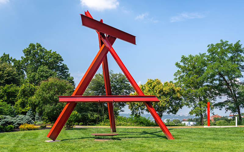 A massive sculpture made from red steel beams. The beams form an A-shape, and a swing hangs in the center