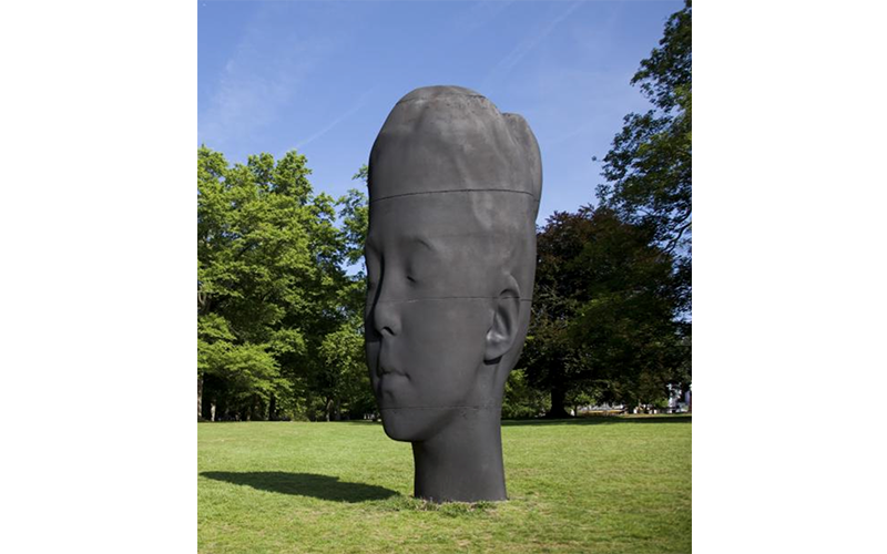 A massive black sculpture of a woman's head with closed eyes. The sculpture is very thin, but looks 3D anyway