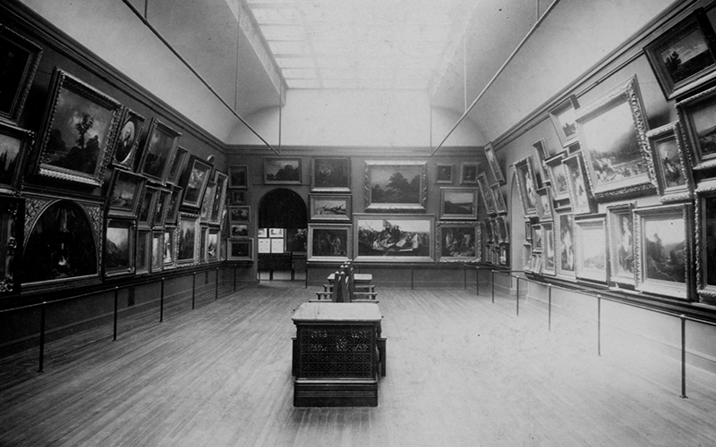 A historic black and white photo of a gallery filled with paintings