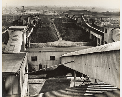 A black and white photo of a coal mining operation, featuring buildings, chutes, and piles of coal