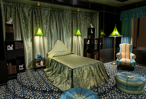 A room with a shiny green bedspread and curtain and a floral carpet