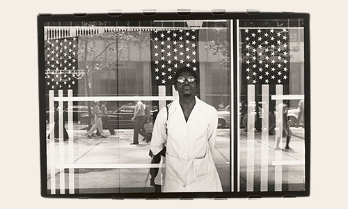A black and white photo of a Black person standing in front of a window with hanging American flags