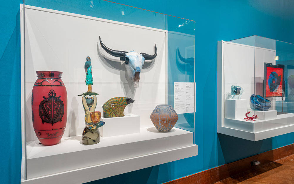 A case filled with colorful artworks including a large red vase with a turtle decoration and a large cow skull with blue shading and dark horns.