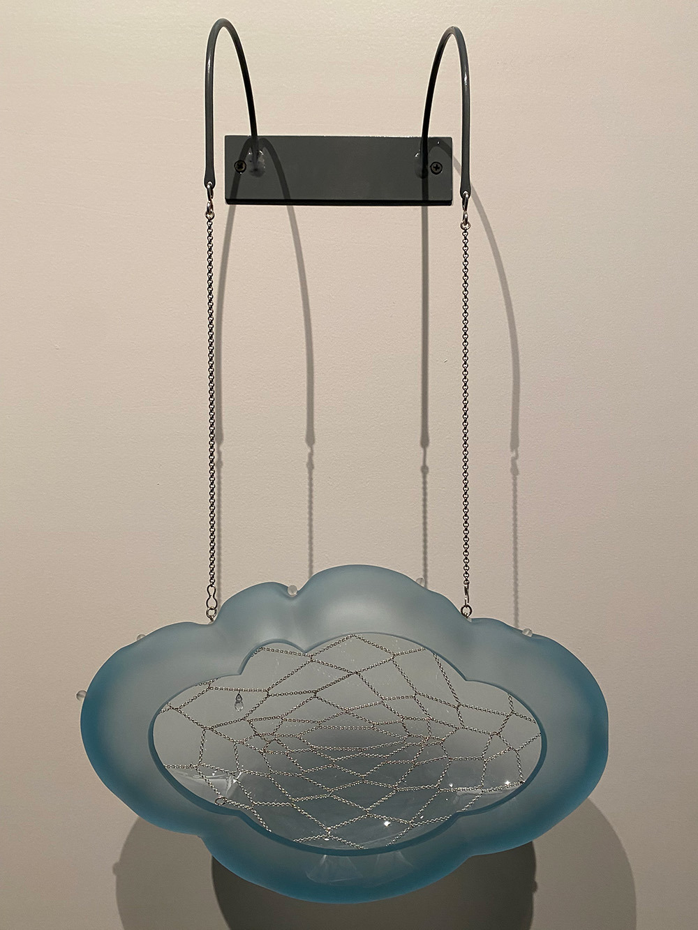 Dream Cloud is made from cut and hand-sculpted glass, silver, and steel. A blue glass cloud is suspended out from the wall by two chains which are attached to a steel wall mount.