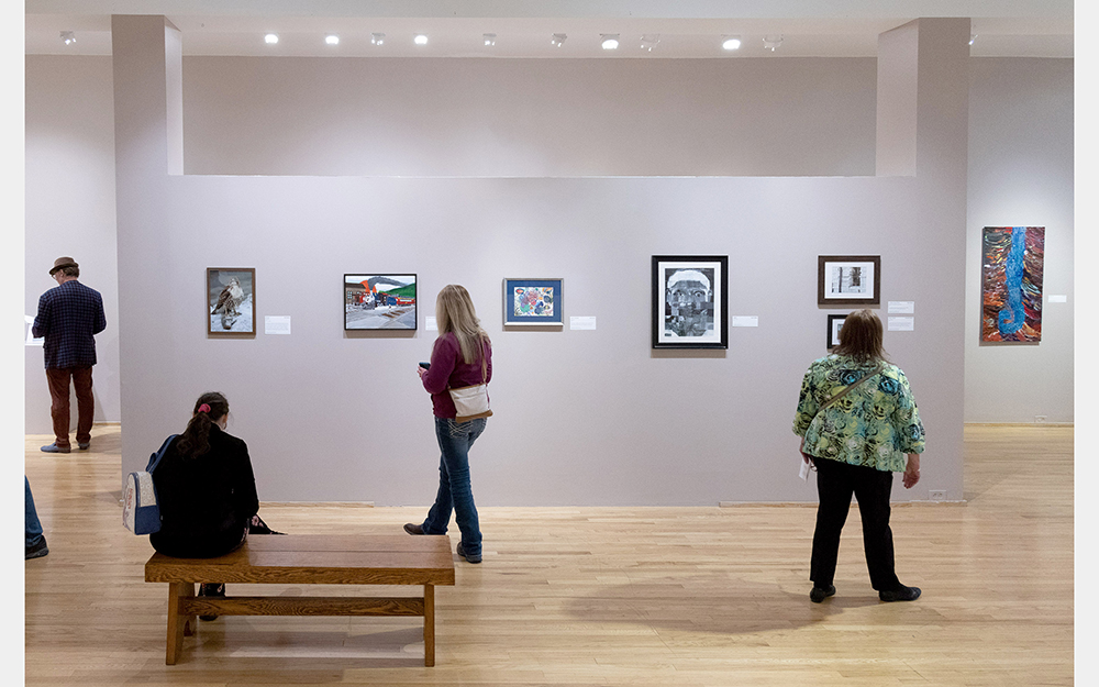 Several people look at art on the walls of a gallery. One person sits on a bench.