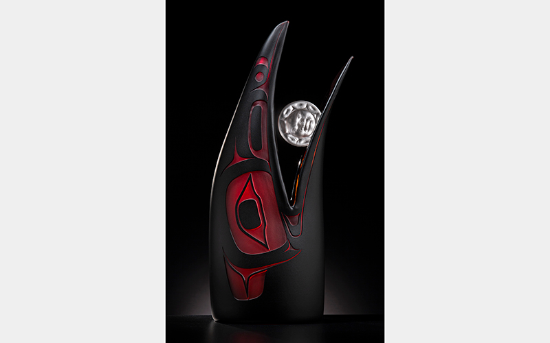 A dark bird-like sculpture with red details rises up and holds a silver circle in its open mouth.