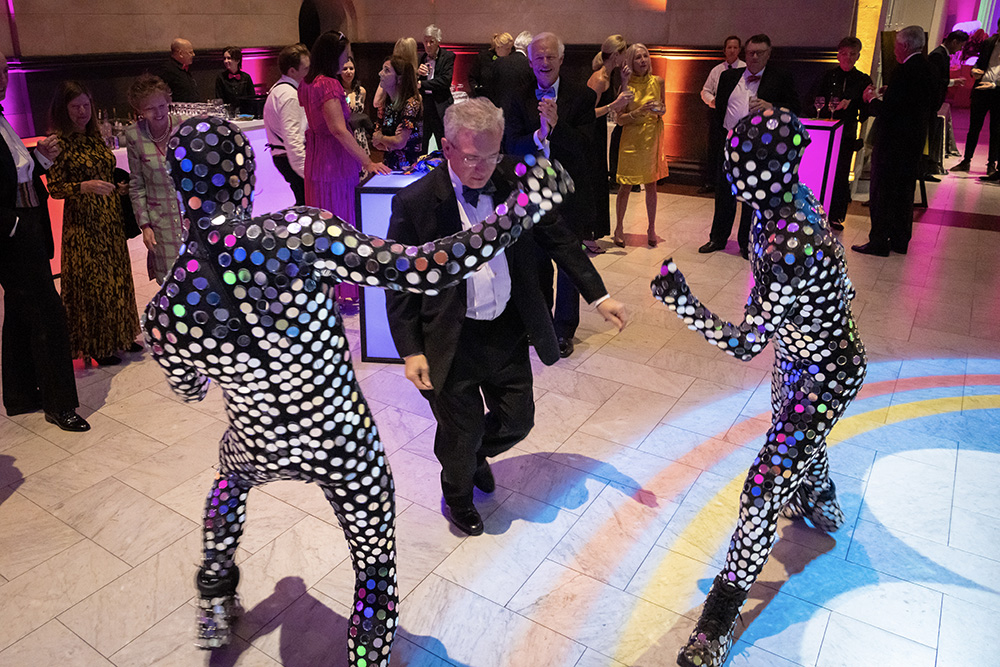 Two people in black suits covered with tiny round mirrors dance with a visitor