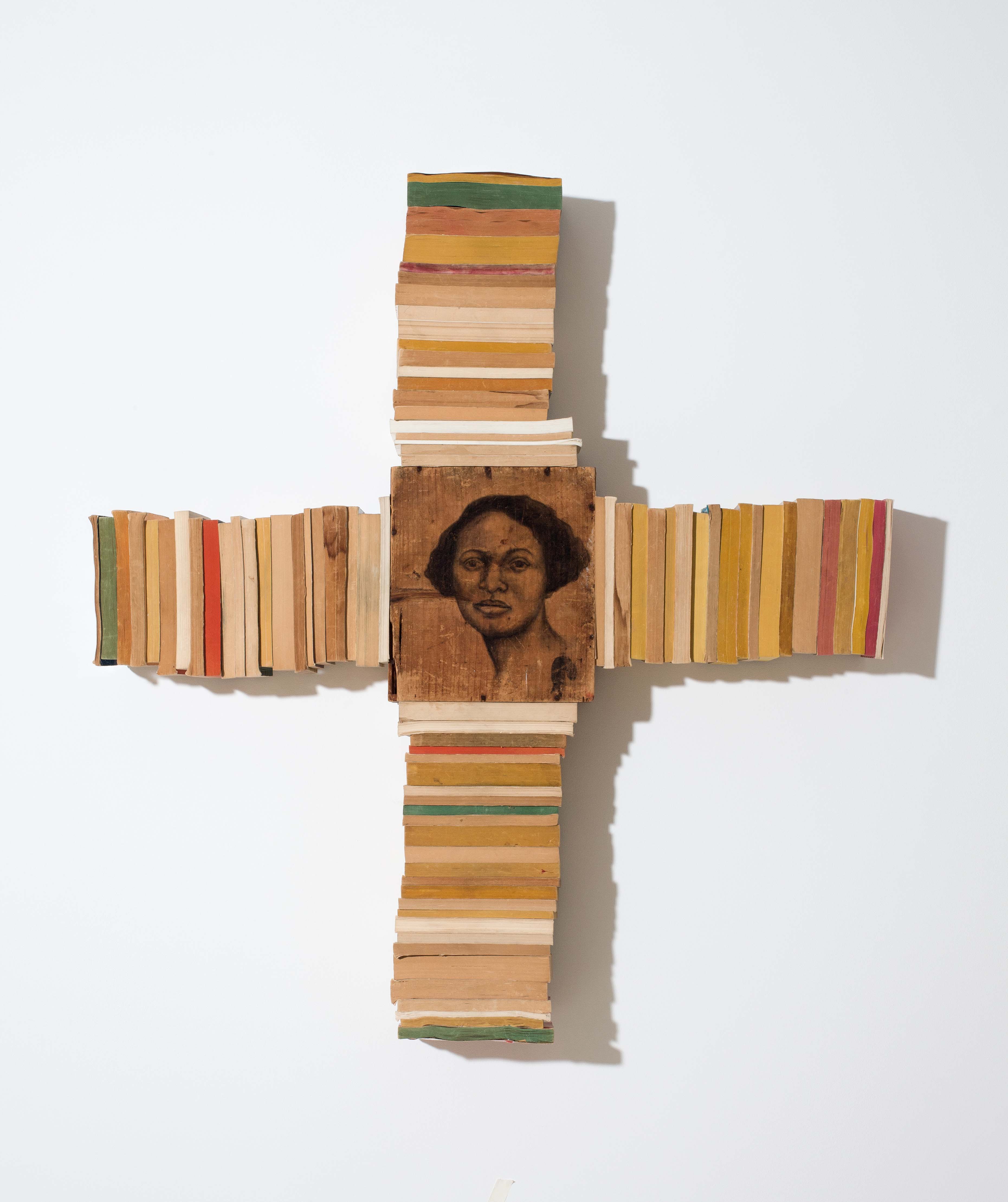 An assortment of books photographed in domino formation (tips up) arranged in a cross formation, a wood panel placed in the center with a woman's face painted with black ink.