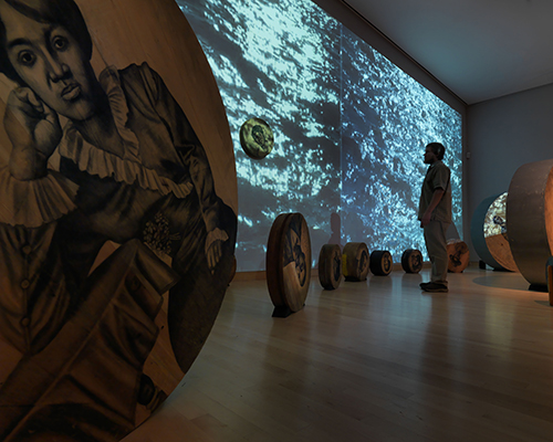 A person stands in a room filled with wooden circles and a projection of moving water. The wooden circles have monochromatic portraits of people on them.