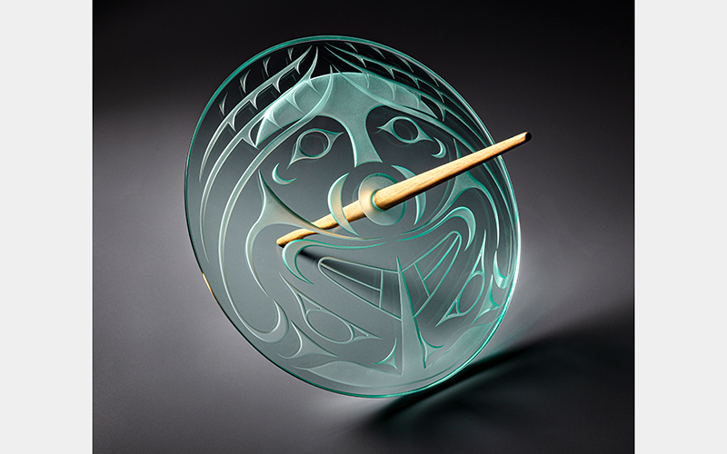 A clear glass disc with a face motif and a thin wooden rod running through it