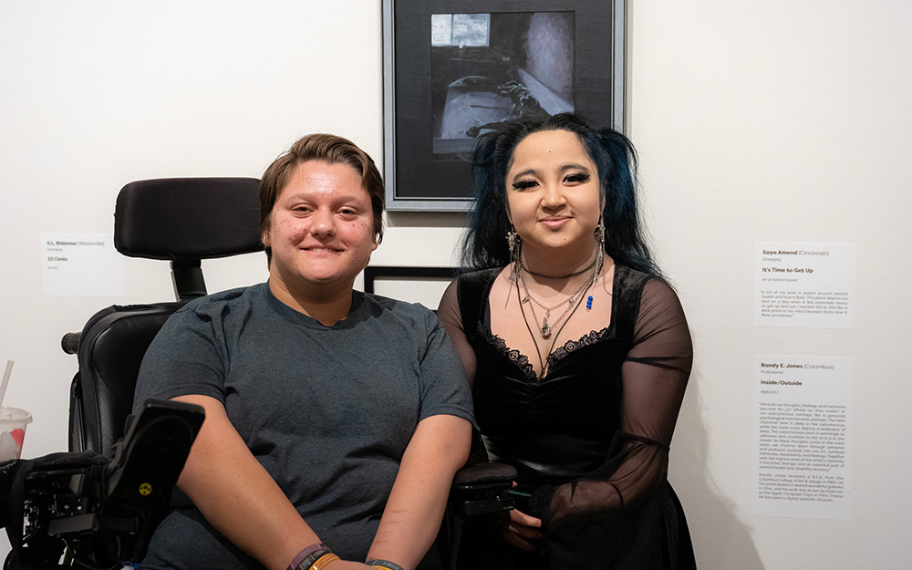 Two people with light skin smile in front of an artwork on a wall. One person sits in a power wheelchair.