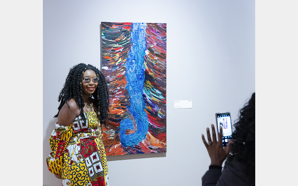A Black woman smiles and gets her picture taken beside a large painting with a blue swirl in the center and multicolored lines surrounding it.
