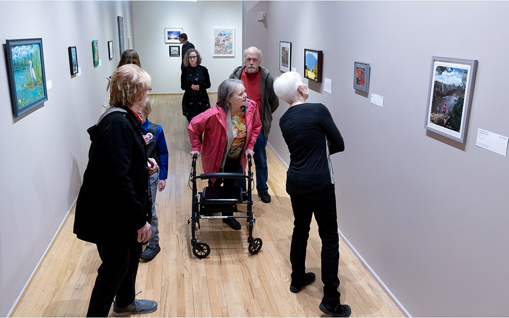 A group of people look at artworks on the walls of a gallery. One woman uses a mobility aid.