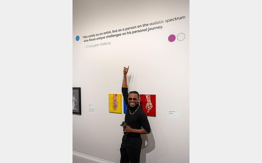A smiling Black man points at a quote on a wall above him. The quote reads "Not solely as an artist, but as a person on the autistic spectrum who faces unique challenges on his personal journey."