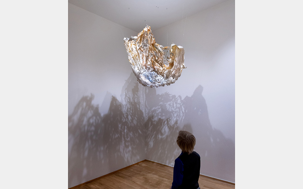A young person looks up at a fluffy, light-colored installation that casts complex shadows with layered, crooked lines.
