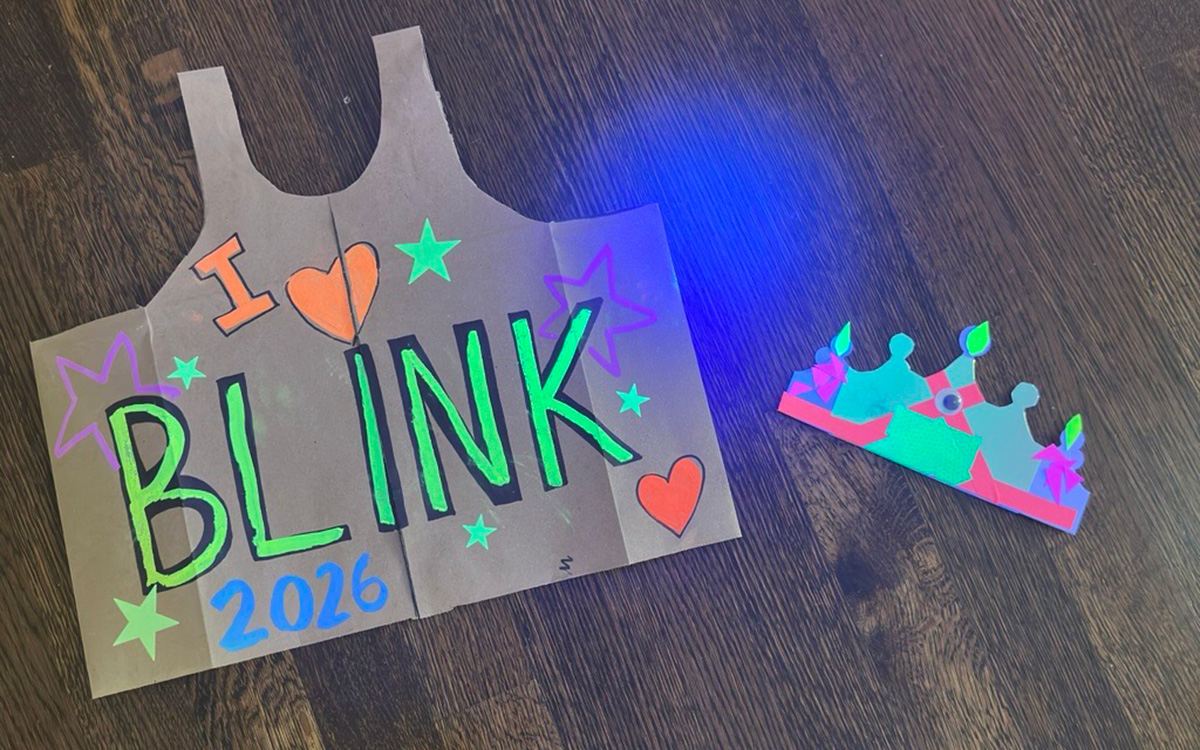 Paper vest and crown decorated with fluorescent paints with a wood flooring background. "I heart BLINK 2026" is written on the vest. 