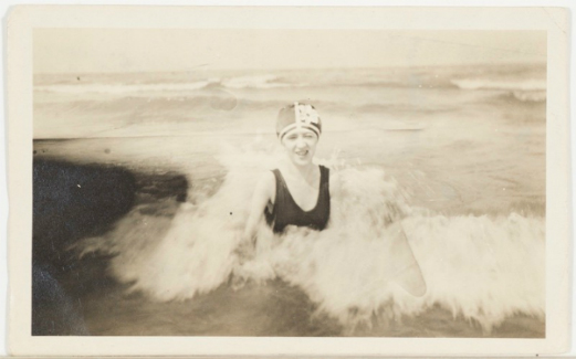 Unidentified Artist, Woman Wearing Flowered Bath-ing Cap in Rushing Surf, black and white photograph of a woman sitting amidst a small wave