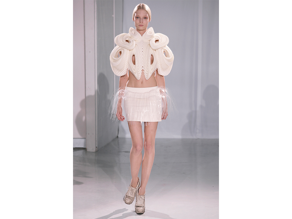Iris van Herpen's Crystallization, an off white mini skirt with transparent string hanging down at the waist, and a large matching top with a feather like texture covering the shoulders and down to the models stomach, made up of several oval shapes