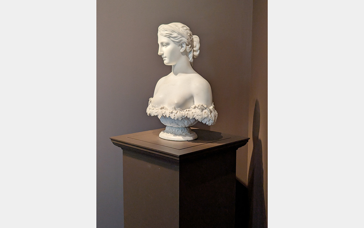A white marble bust of a woman sits on a brown pedestal against a light brown background.