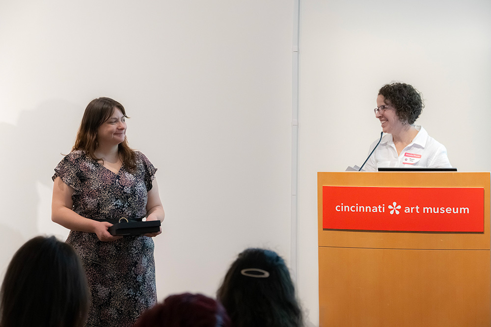 Sara Birkofer holds an award and smiles at a presenter at a podium