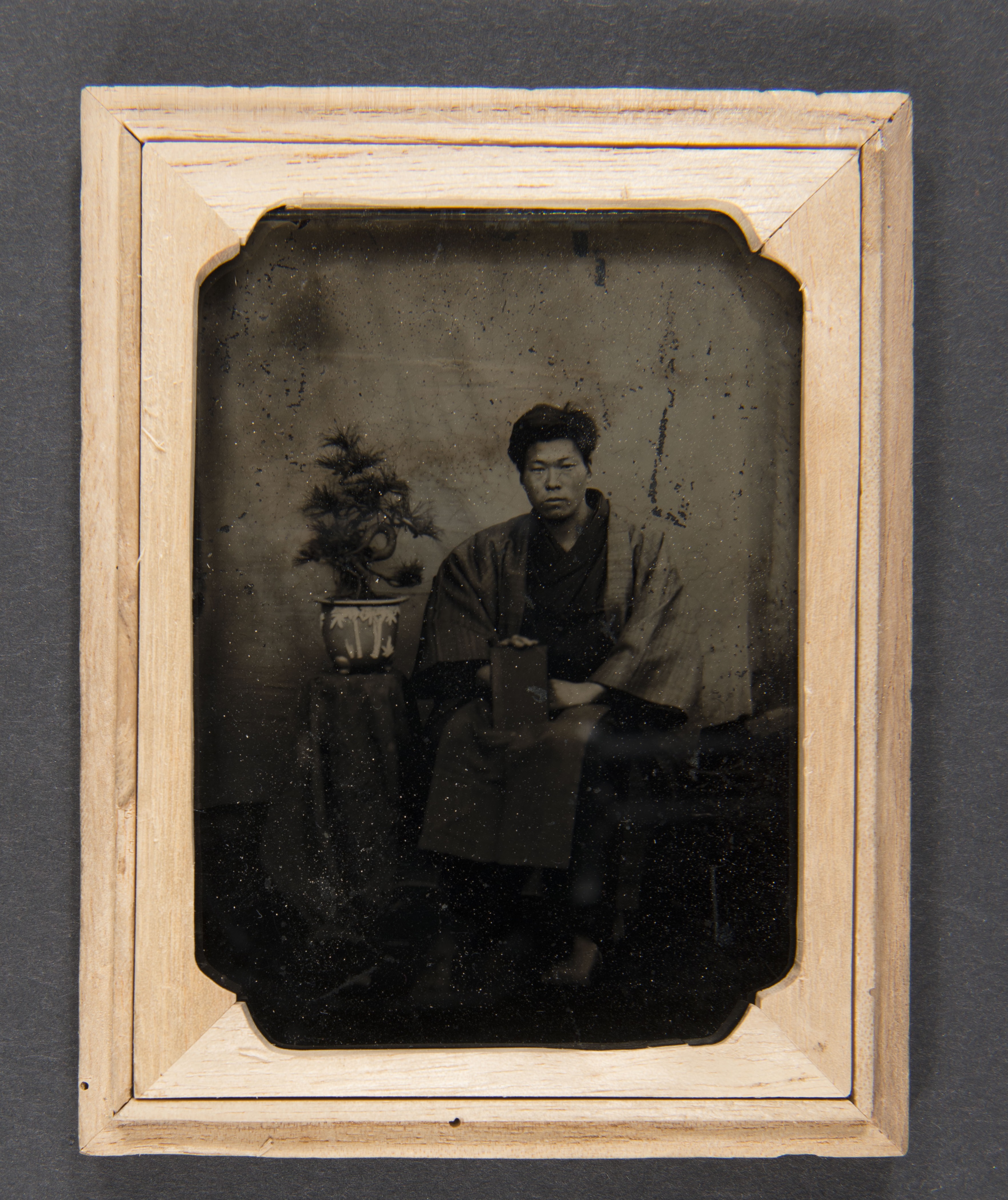 Black and white photograph of a Japanese man sitting next to an end table with a bonsai plant