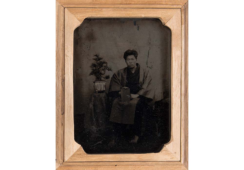 Black and white photograph of a Japanese man sitting next to an end table with a bonsai plant