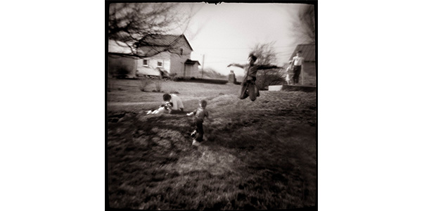 Nancy Rexroth's Boys Flying, a blurry black and white photograph of boys playing in a field. One boy leaps into the air with his arms outstretched to the sides like wings