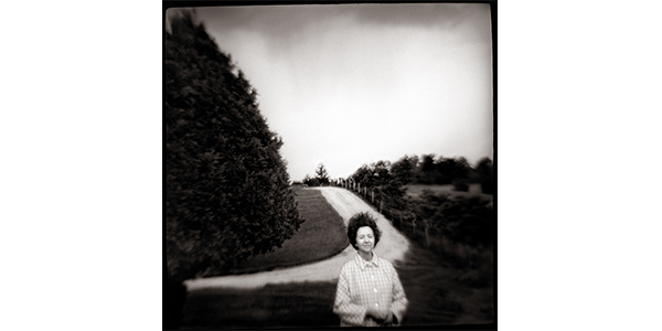 Nancy Rexroth's My Mother, black and white photograph of a woman standing in front of a dirt road curving around a large evergreen tree