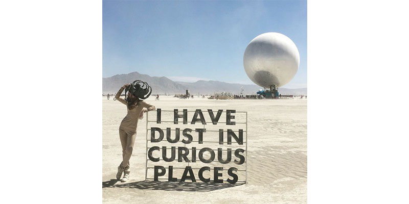 Photograph of a woman in a flat, desert landscape leaning against a sign that says: I have dust in curious places. Behind her is a huge ominous orb. Mountains can be seen in the distance