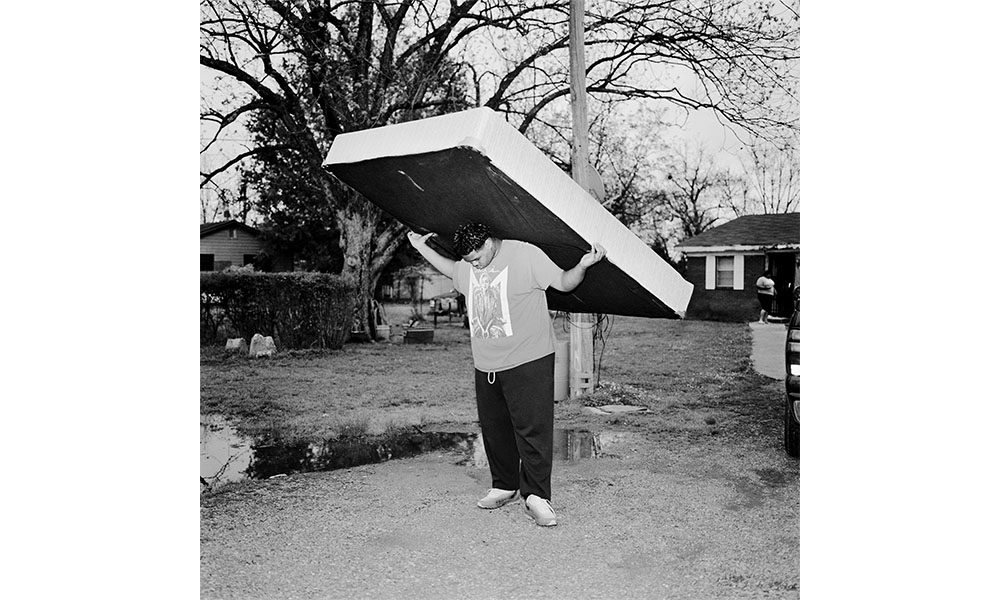 Black and white photograph of an African American boy holding a mattress on his back