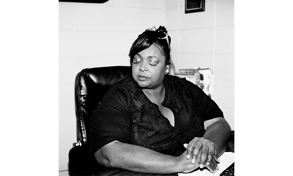 Black and white photograph of an African American woman sitting in a leather chair at a desk. Her eyes are closed and she cups her right hand into her left on the desk