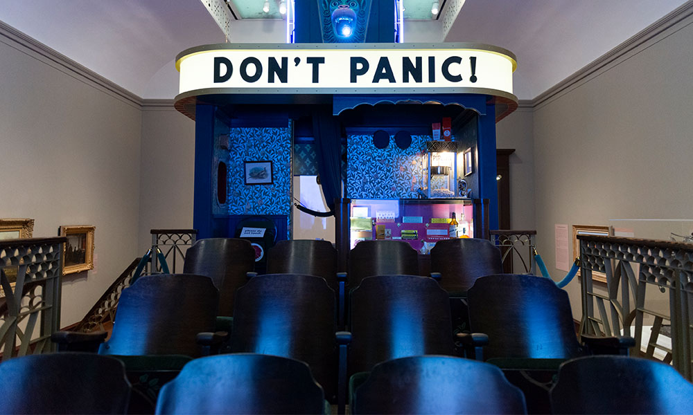 An installation in the exhibition, a lit sign reads: Don't' Panic, above a few rows of seats