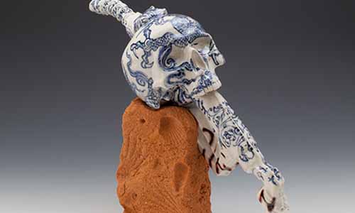 a porcelain statue of a skull embedded onto a fuel nozzle, decorated with a Chinese style dragon in blue glaze. The skull and nozzle rest on a brick impressed with fossilized shells