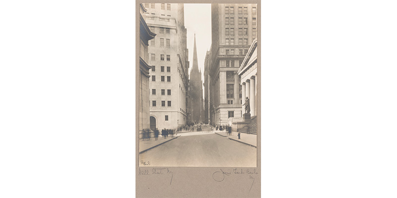Jessie Tarbox Beals' Wall Street, N.Y., an old black and white photograph of Wall Street. Towering skyscrapers dwarf the pedestrians walking around. A hazy spire from a church can been seen in the distance