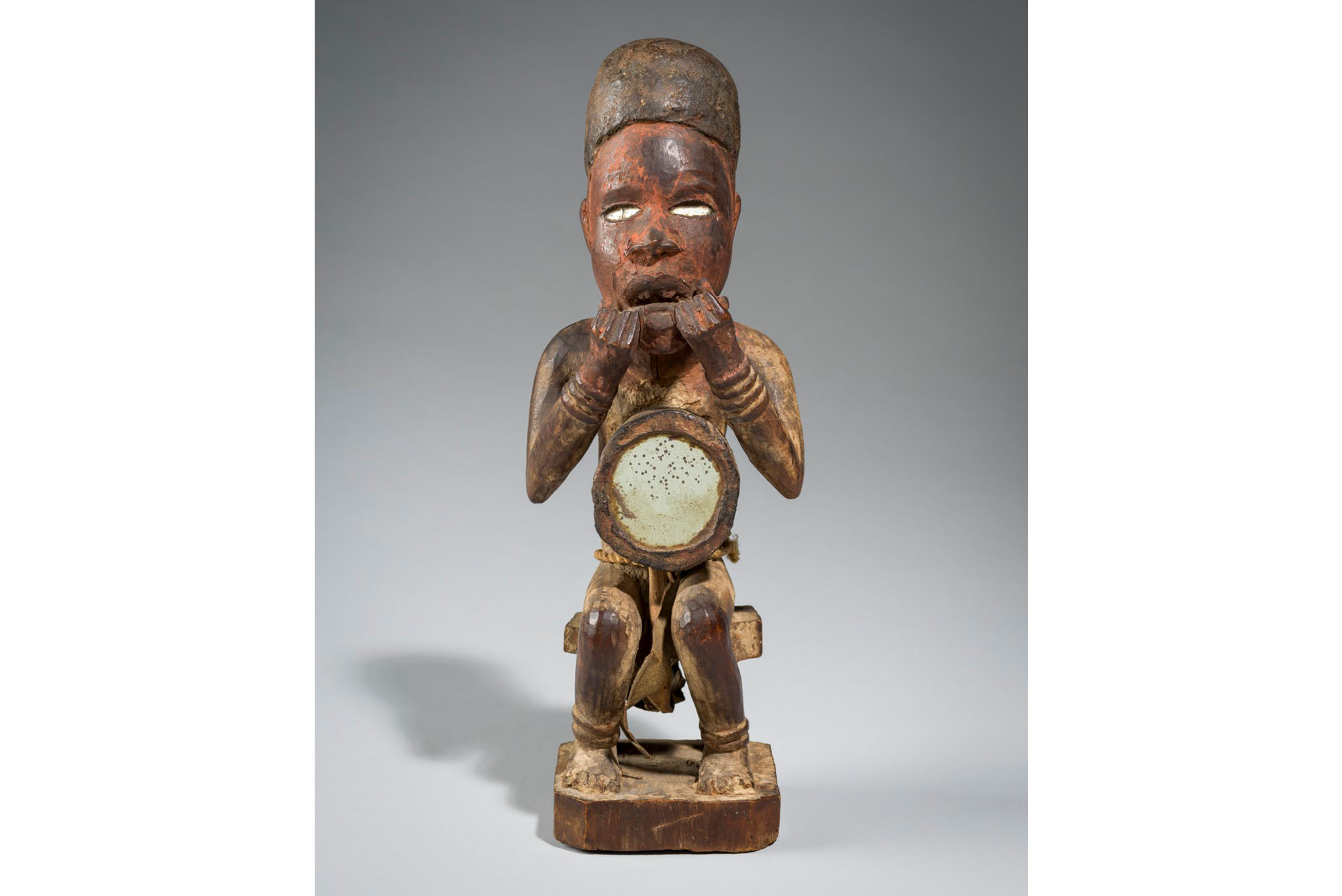 Male Nkisi Power figure, a wooden stature of an African man sitting down with his hands held up to his mouth. In his lap is a small, circular mirror