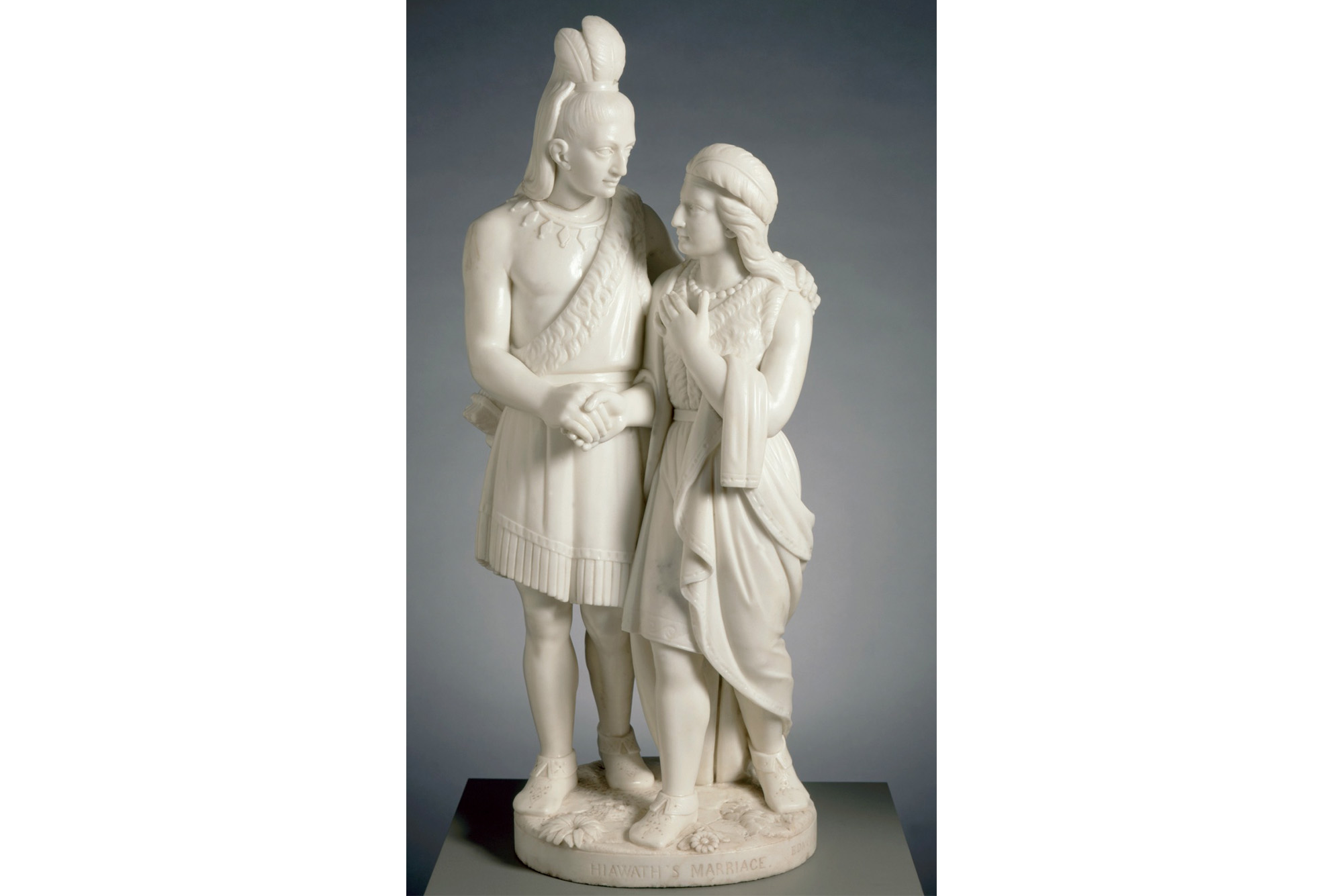 Edmonia Lewis' Hiawatha's Marriage, a marble sculpture of a Native American man and woman. The woman stands slightly ahead of the man and hold his right hand in hers, placing her left hand on her chest. The man places his left hand on her left shoulder as they look towards each other.