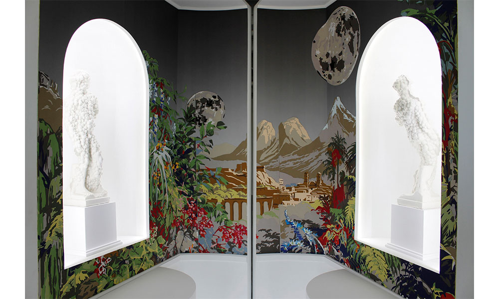 Image of Order, two statues of human figures are posed in large windows on the left and right facing each other. Surround the statues is hand cut paper depicting a lush forest of ferns, flowers and trees. In the distance, a city sits beneath a mountain range lit by two large moons