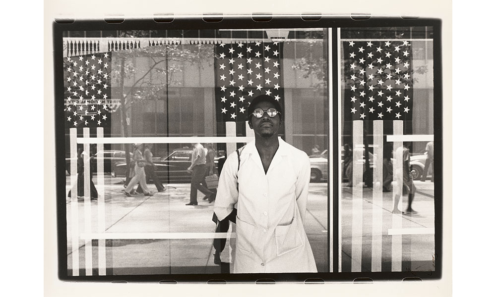 Ming Smith's America seen through Stars and Stripes, black and white photograph of an African American man standing in front of a window reflecting the New York City sidewalk, behind the window hang three American flag banners