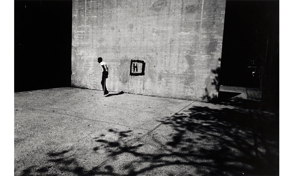 Louis Draper's Untitled [Boy and H], black and white photograph of a lone African American boy sauntering away from an "H" written in a black square on the wall behind him