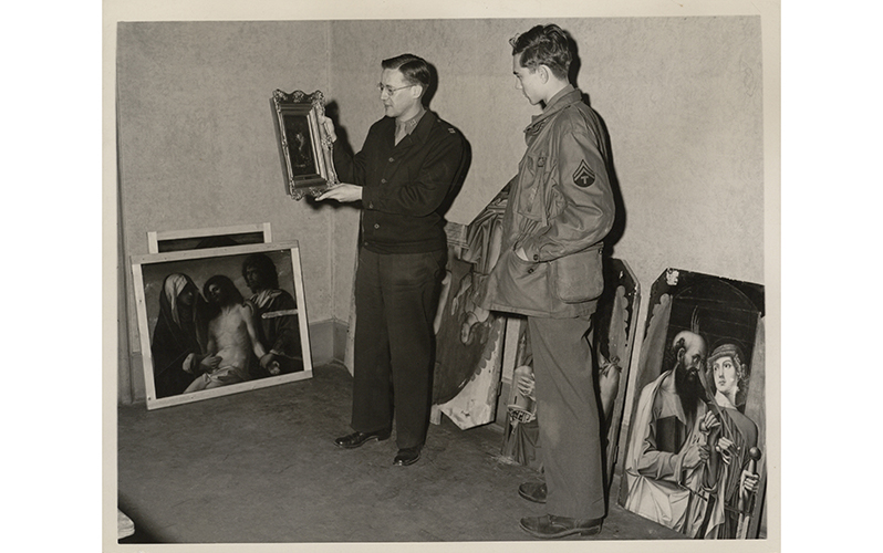 A photograph of two men in a room with a collection of art work leaning against the wall. The man on the left shows a painting to the other man
