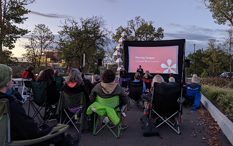 Visitors sit outside in camp chairs watching a speaker give a presentation