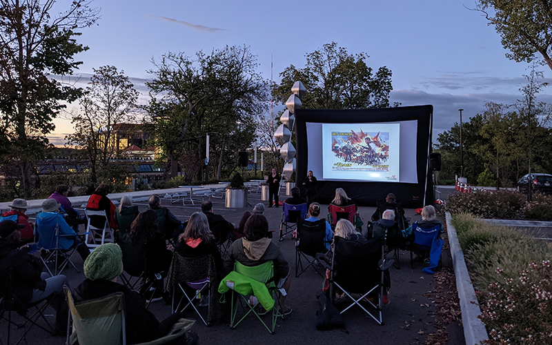 Visitors sit outside in camp chairs watching speakers give a presentation