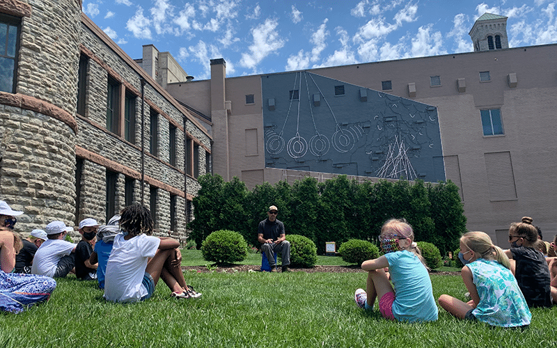 A group of kids sit in the grass and listen to a presentation