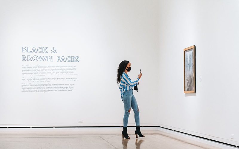 A woman of color wearing a mask takes a picture of an artwork hanging on a wall. "Black & Brown Faces" is visible on the wall behind her.