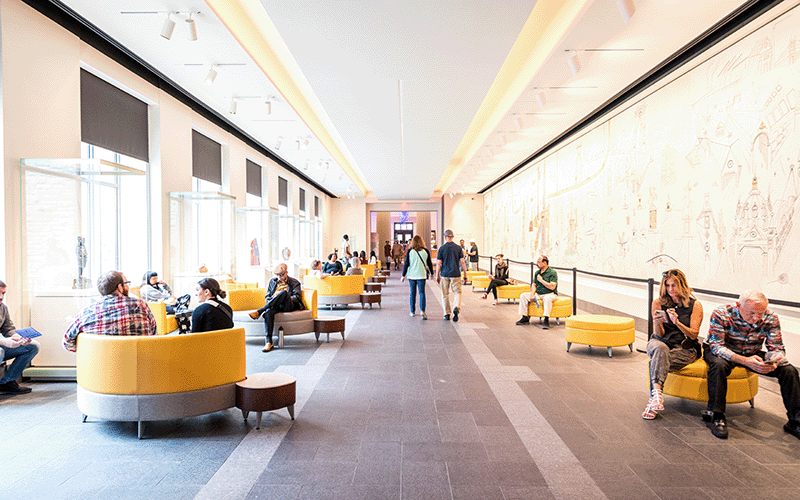 A long hallway with yellow furniture is filled with sitting people