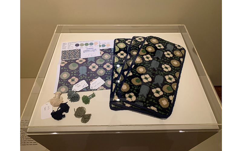 A recreated design of Elaine Wormser’s carpet on paper, next to woven carpet samples and colored yarns in beige, grass green, olive green, and dark and light blue. 