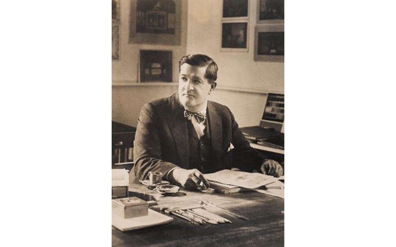 Joseph Urban seated at his desk, looking away from the camera. Framed pictures hang on the wall behind him.