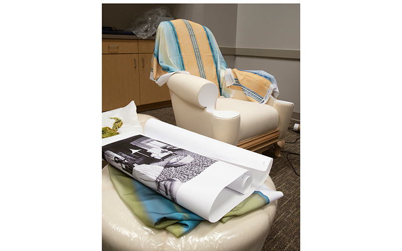Reproduction of Elaine Wormser’s armchair, with blue and yellow striped sample upholstery draped over it. In the foreground, lying on top of a reproduction hassock, is a printout of the black and white photograph of the chair and a hassock. 