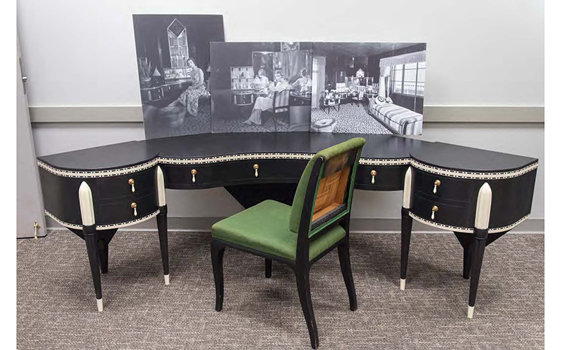 A curved dressing table and chair, with enlarged archival photos of Elaine Wormser’s bedroom sitting on the table.  